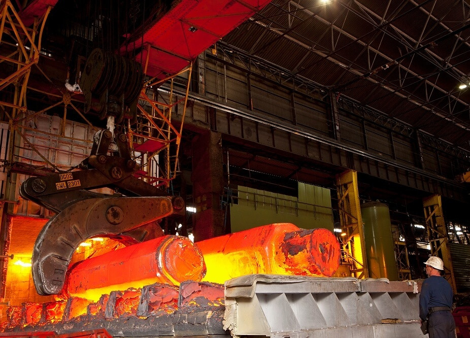 Worker in a steel mill observing large glowing hot metal pieces being moved by an overhead crane.